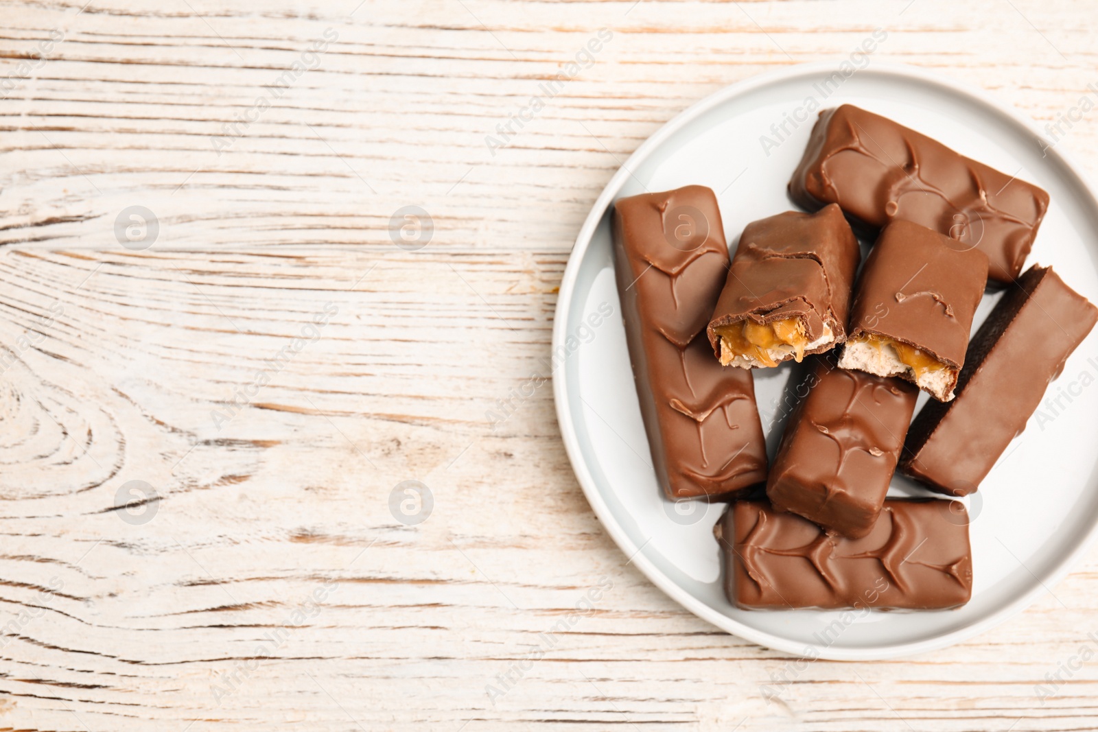 Photo of Plate of chocolate bars with caramel, nuts and nougat on white wooden table, top view. Space for text