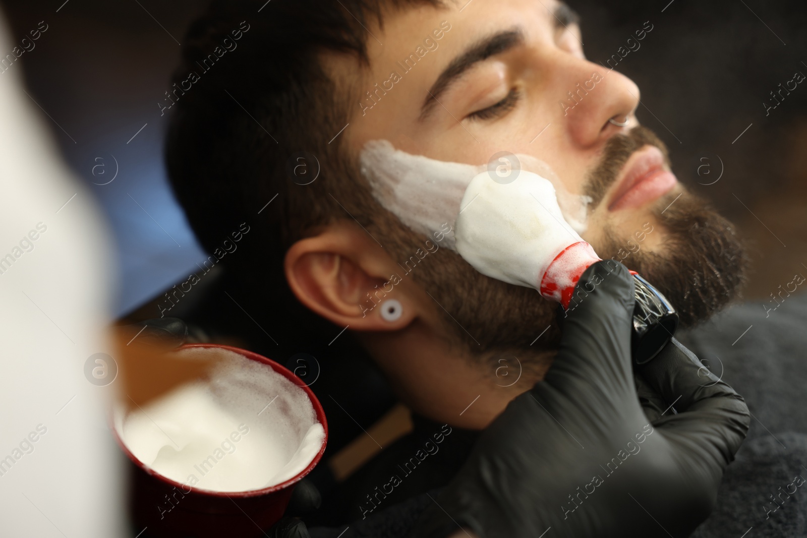 Photo of Professional hairdresser applying shaving foam onto client's beard in barbershop, closeup