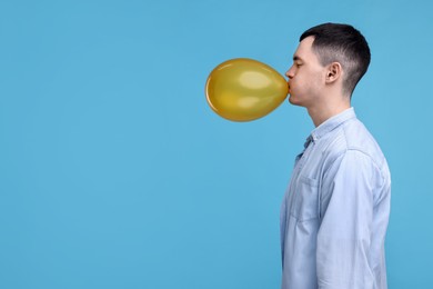 Photo of Young man inflating golden balloon on light blue background. Space for text