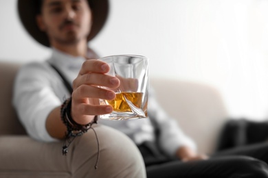 Young man with glass of whiskey sitting on sofa at home. Space for text