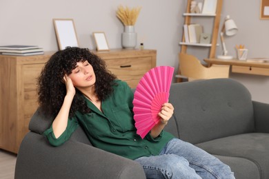 Photo of Young woman waving pink hand fan to cool herself on sofa at home. Space for text