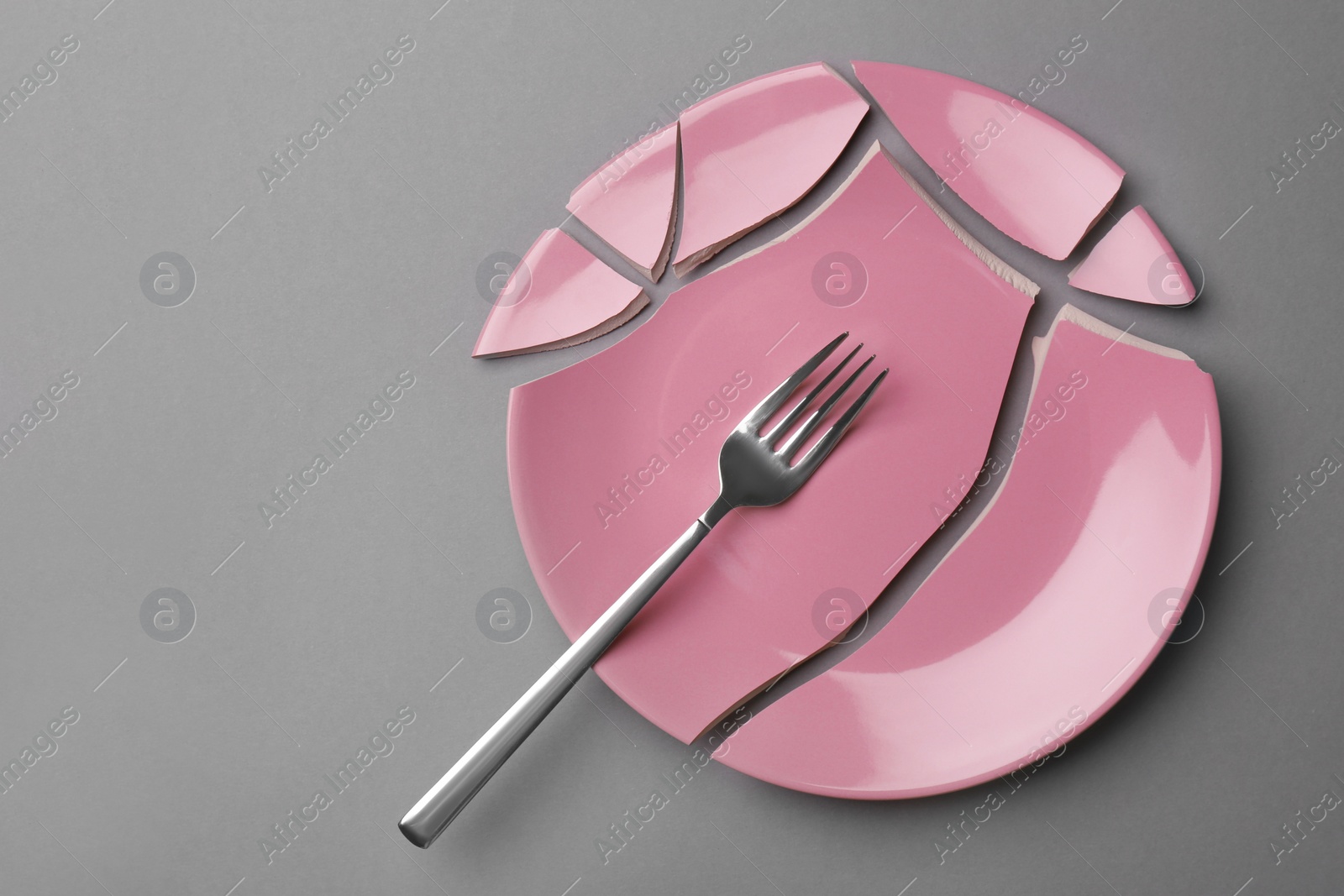 Photo of Pieces of broken pink ceramic plate and fork on grey background, top view. Space for text