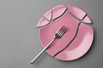 Pieces of broken pink ceramic plate and fork on grey background, top view. Space for text