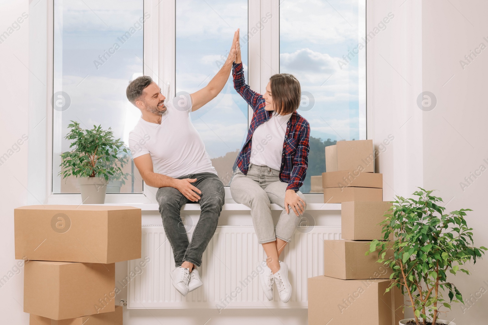 Photo of Happy couple giving high five while sitting on windowsill in new apartment. Moving day