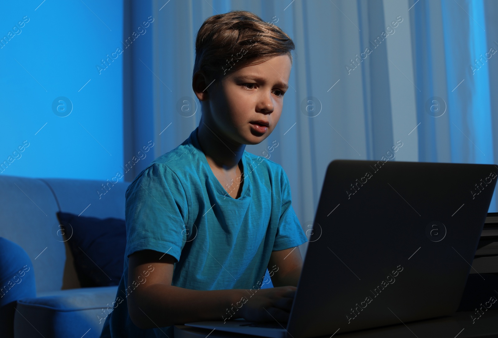 Photo of Shocked little child with laptop in dark room. Danger of internet