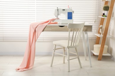 Photo of White sewing machine and fabric on wooden desk near chair indoors