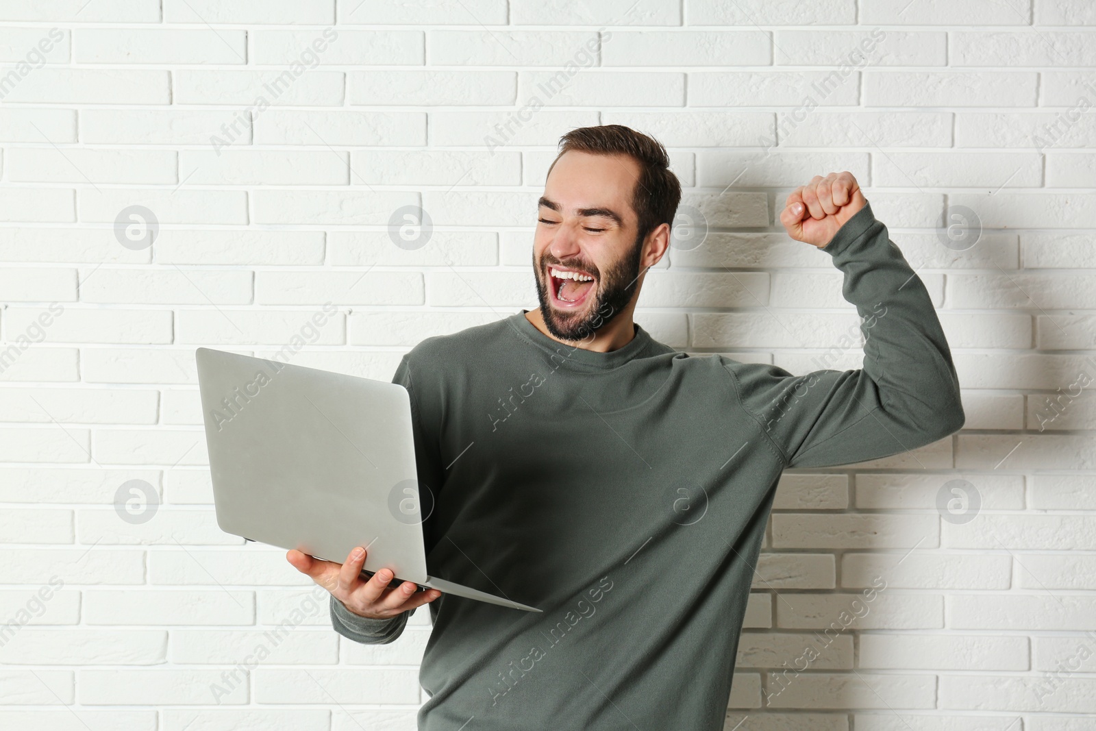 Photo of Emotional young man with laptop celebrating victory near brick wall