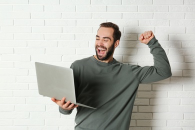 Photo of Emotional young man with laptop celebrating victory near brick wall