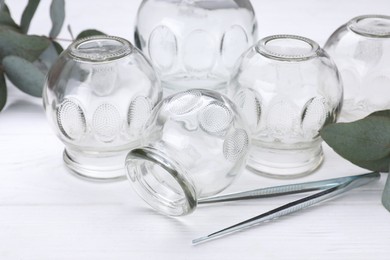 Photo of Glass cups, tweezers and eucalyptus leaves on white wooden table, closeup. Cupping therapy