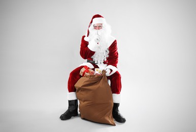 Photo of Santa Claus sitting near sack with gift boxes on light grey background