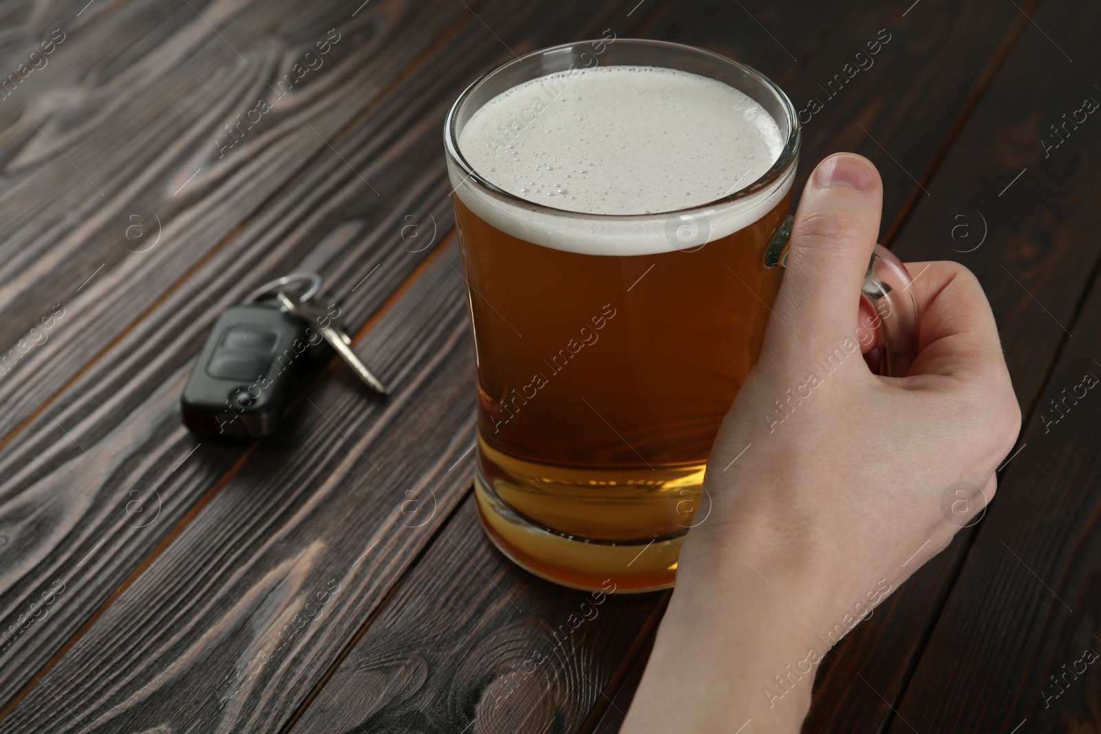 Photo of Man holding glass of alcoholic beer near car key at wooden table, closeup. Dangerous drinking and driving