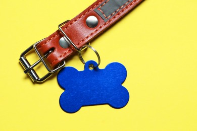 Photo of Leather dog collar with blue tag in shape of bone on yellow background, top view. Space for text