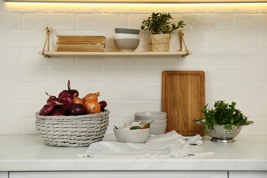 Photo of Fresh onions in basket and different kitchenware on countertop indoors