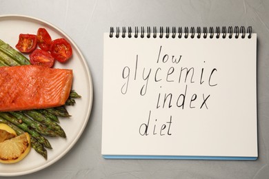 Notebook with words Low Glycemic Index Diet and plate of tasty grilled salmon on grey table, flat lay
