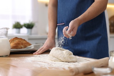 Photo of Making bread. Man sprinkling flour onto dough at wooden table in kitchen, closeup