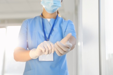 Photo of Doctor in protective mask and scrubs putting on medical gloves indoors, closeup