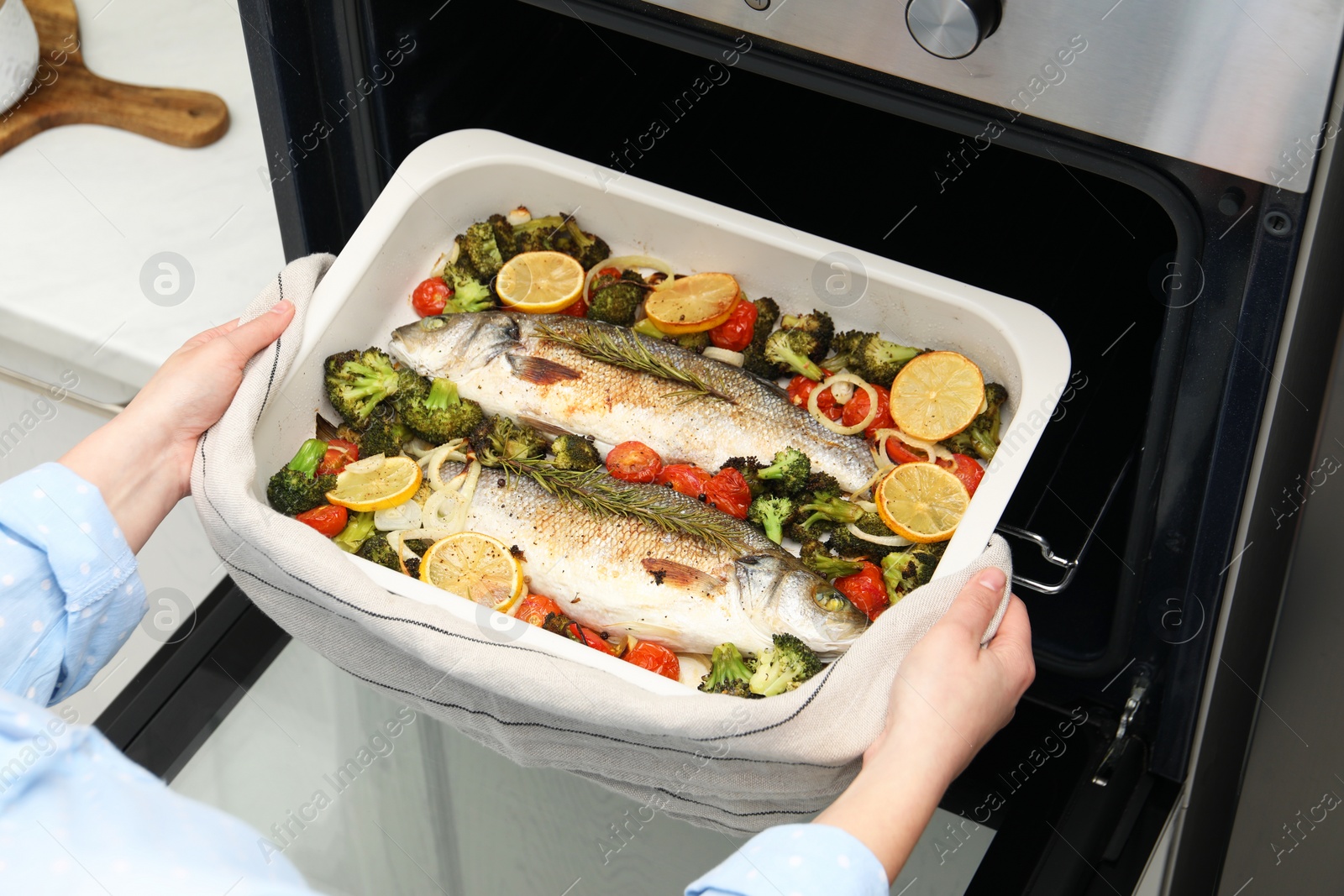 Photo of Woman taking baking dish with delicious fish and vegetables from oven in kitchen, closeup