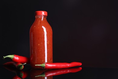 Photo of Spicy chili sauce in bottle and peppers against dark background, space for text