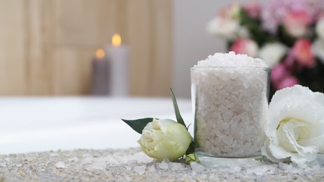 Photo of Glass with bath salt and beautiful flowers on wicker mat in bathroom, closeup. Space for text