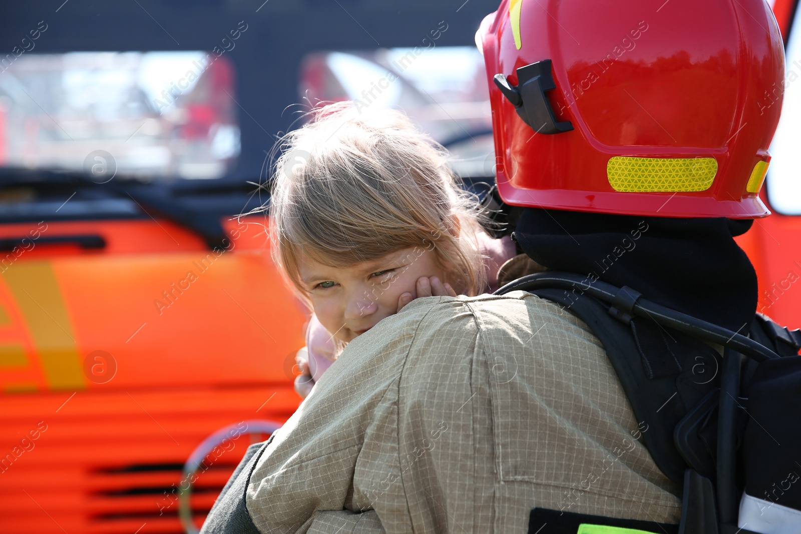 Photo of Firefighter in uniform holding rescued little girl near fire truck outdoors. Save life