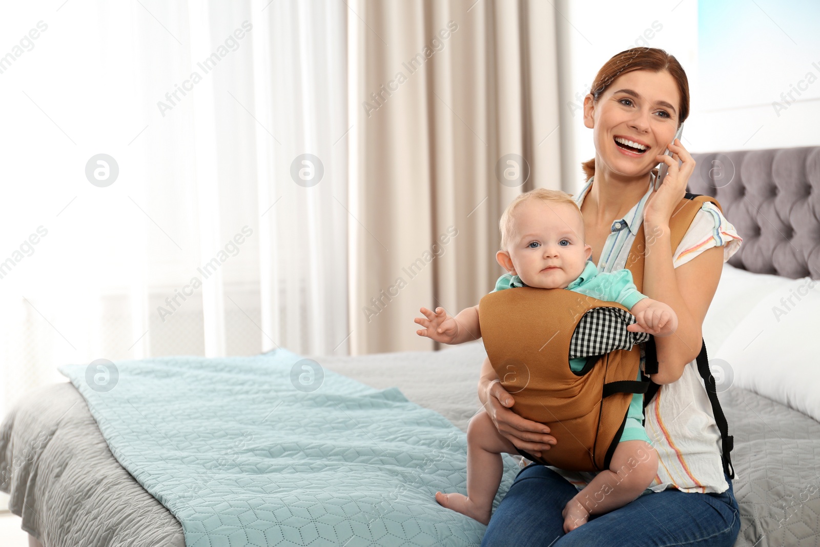 Photo of Woman with her son in baby carrier talking on phone at home. Space for text