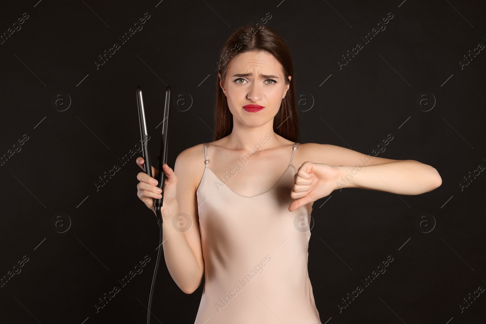 Photo of Upset young woman with flattening iron showing thumb down on black background. Hair damage