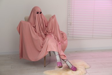 Glamorous ghost. Woman in pink sheet and high heel shoes on armchair indoors, space for text