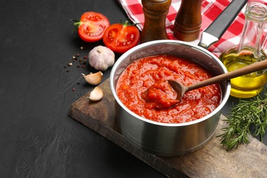 Photo of Homemade tomato sauce in pot, spoon and ingredients on dark table, space for text