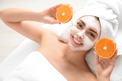 Photo of Pretty woman with rejuvenating facial mask holding sliced orange in spa salon, above view