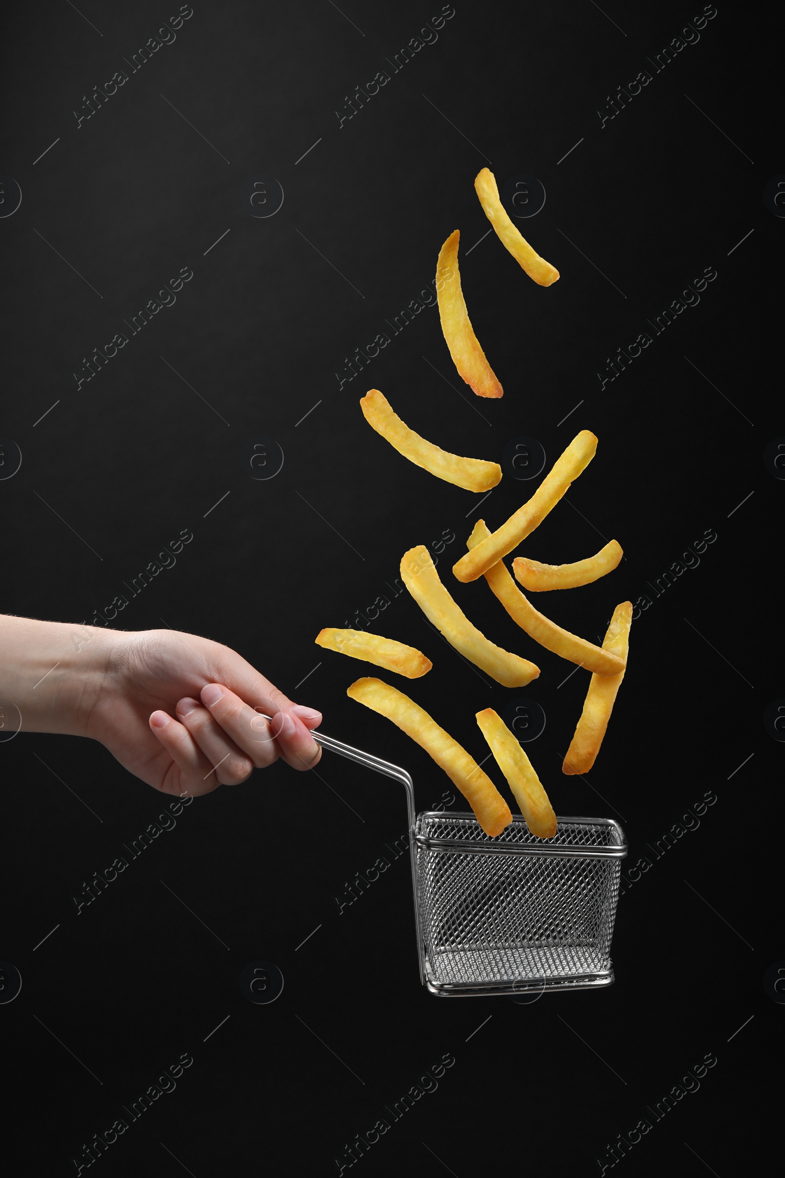 Image of French fries falling into metal basket held by woman on black background, closeup