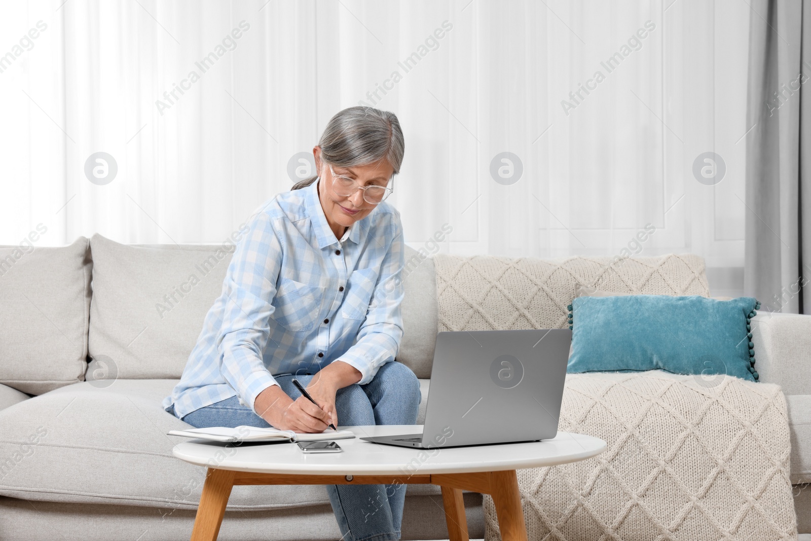 Photo of Beautiful senior woman writing something in notebook while working with laptop at home