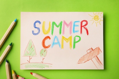 Photo of Paper with written text SUMMER CAMP, drawings and different pencils on color background, flat lay