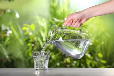 Woman pouring water from jug into glass on light grey table outdoors, closeup