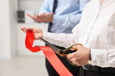 Woman cutting red ribbon with scissors indoors, closeup