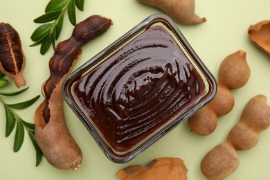 Photo of Bowl of tasty tamarind sauce, leaves and fresh pods on light green table, flat lay