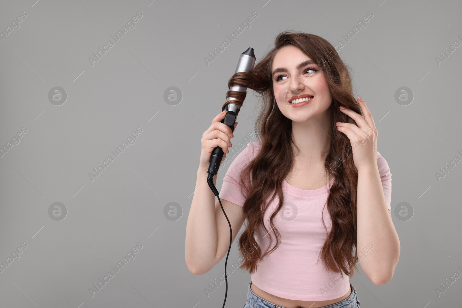 Photo of Beautiful young woman using curling hair iron on grey background, space for text