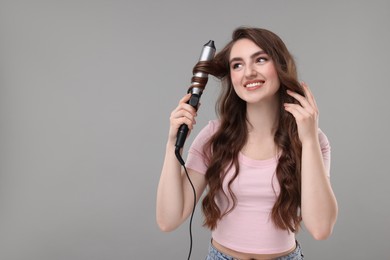 Beautiful young woman using curling hair iron on grey background, space for text