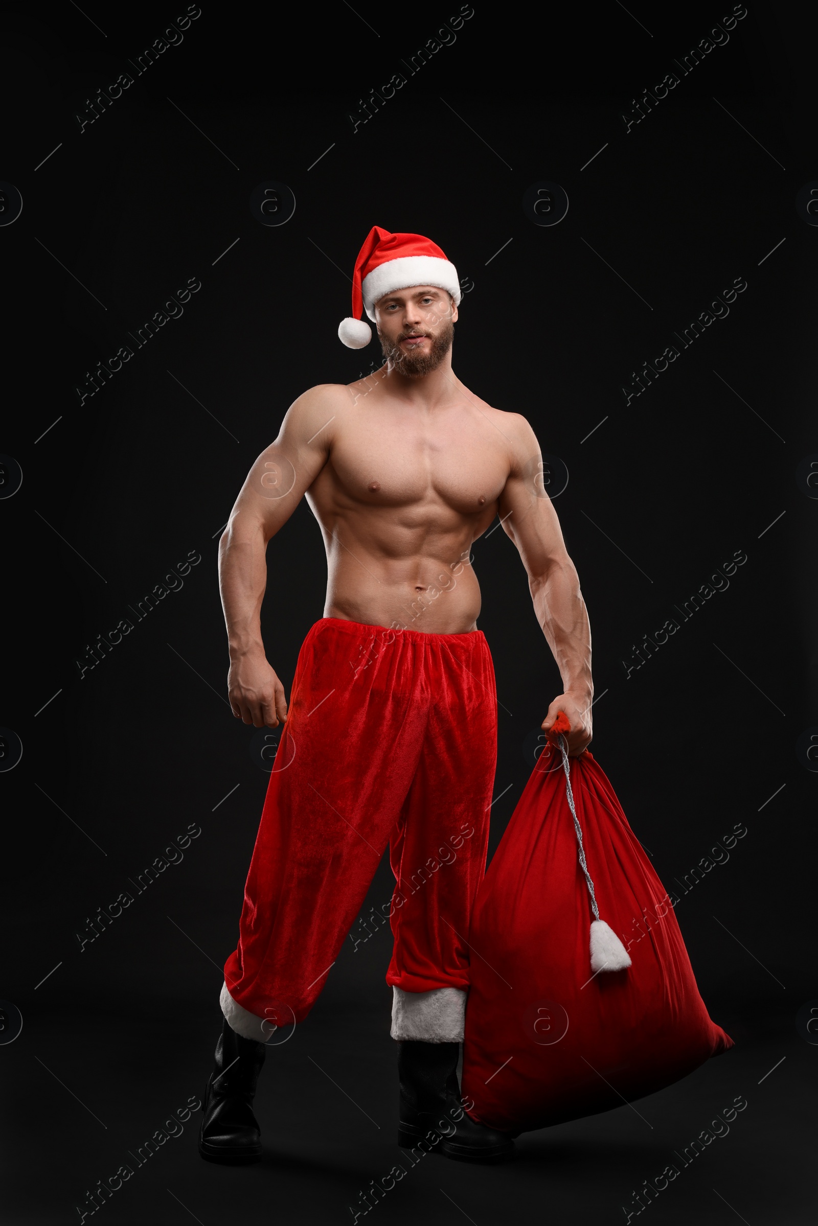 Photo of Muscular young man in Santa hat holding bag with presents on black background