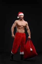 Muscular young man in Santa hat holding bag with presents on black background