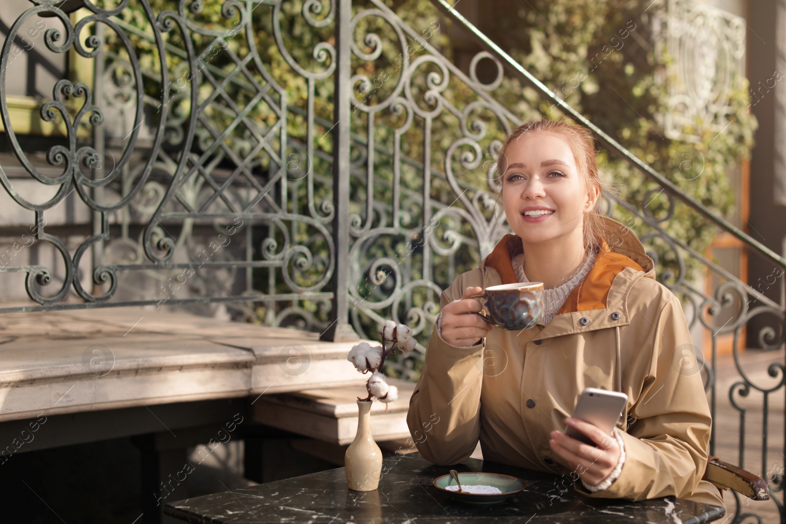 Photo of Young woman enjoying tasty coffee while using mobile phone at table outdoors