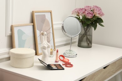 Photo of Mirror, cosmetic products, perfume and vase with pink roses on white dressing table in makeup room