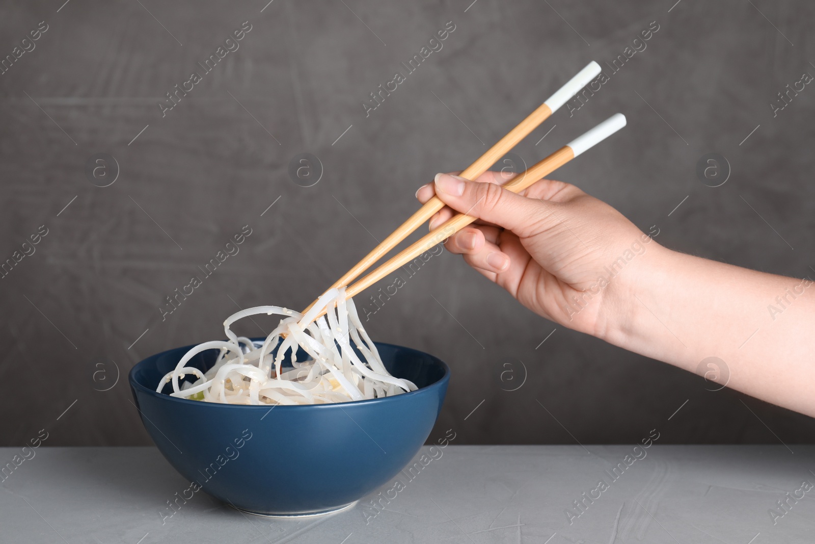 Photo of Woman eating cooked Asian noodles with chopsticks on table against grey background, closeup