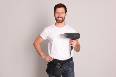 Photo of Smiling hairdresser with tool bag holding dryer on light grey background
