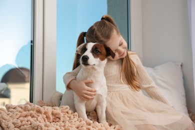 Photo of Cute little girl with her dog sitting on window sill indoors. Childhood pet