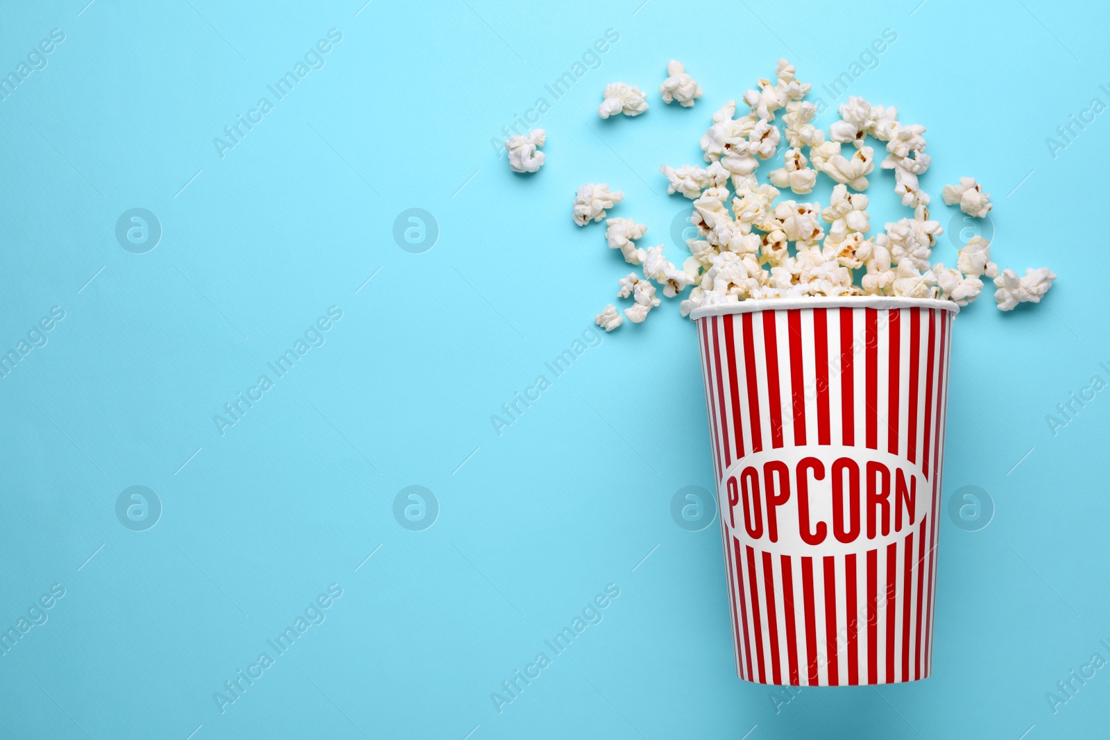 Photo of Overturned paper cup with delicious popcorn on light blue background, flat lay. Space for text