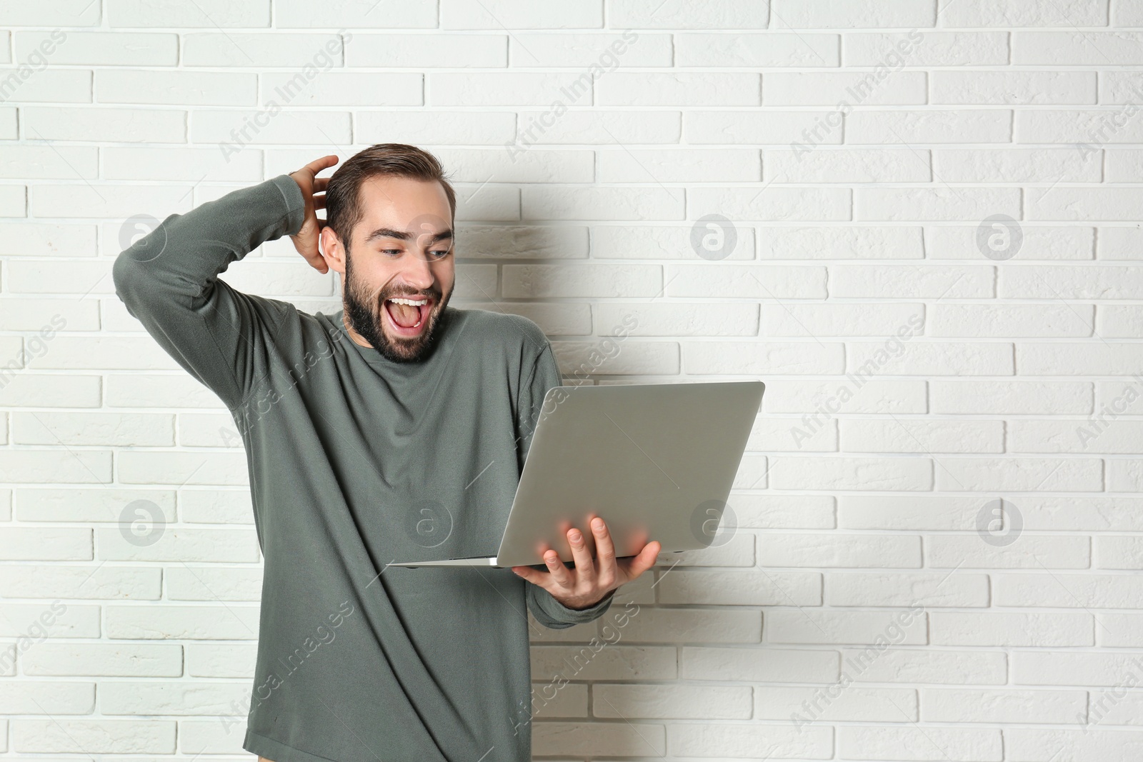 Photo of Emotional young man with laptop celebrating victory near brick wall. Space for text