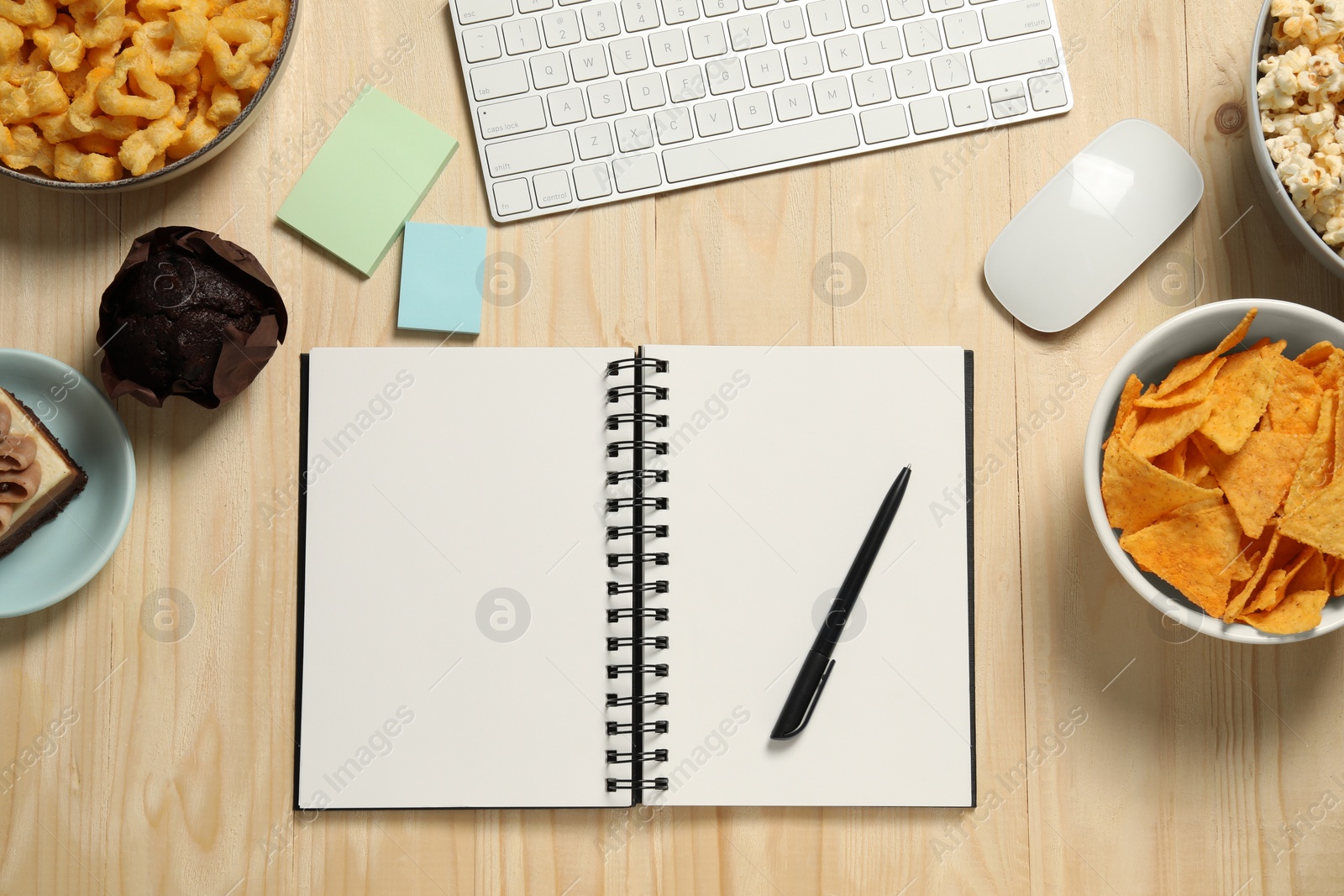 Photo of Bad eating habits at workplace. Keyboard, notebook and different snacks on wooden table, flat lay. Space for text