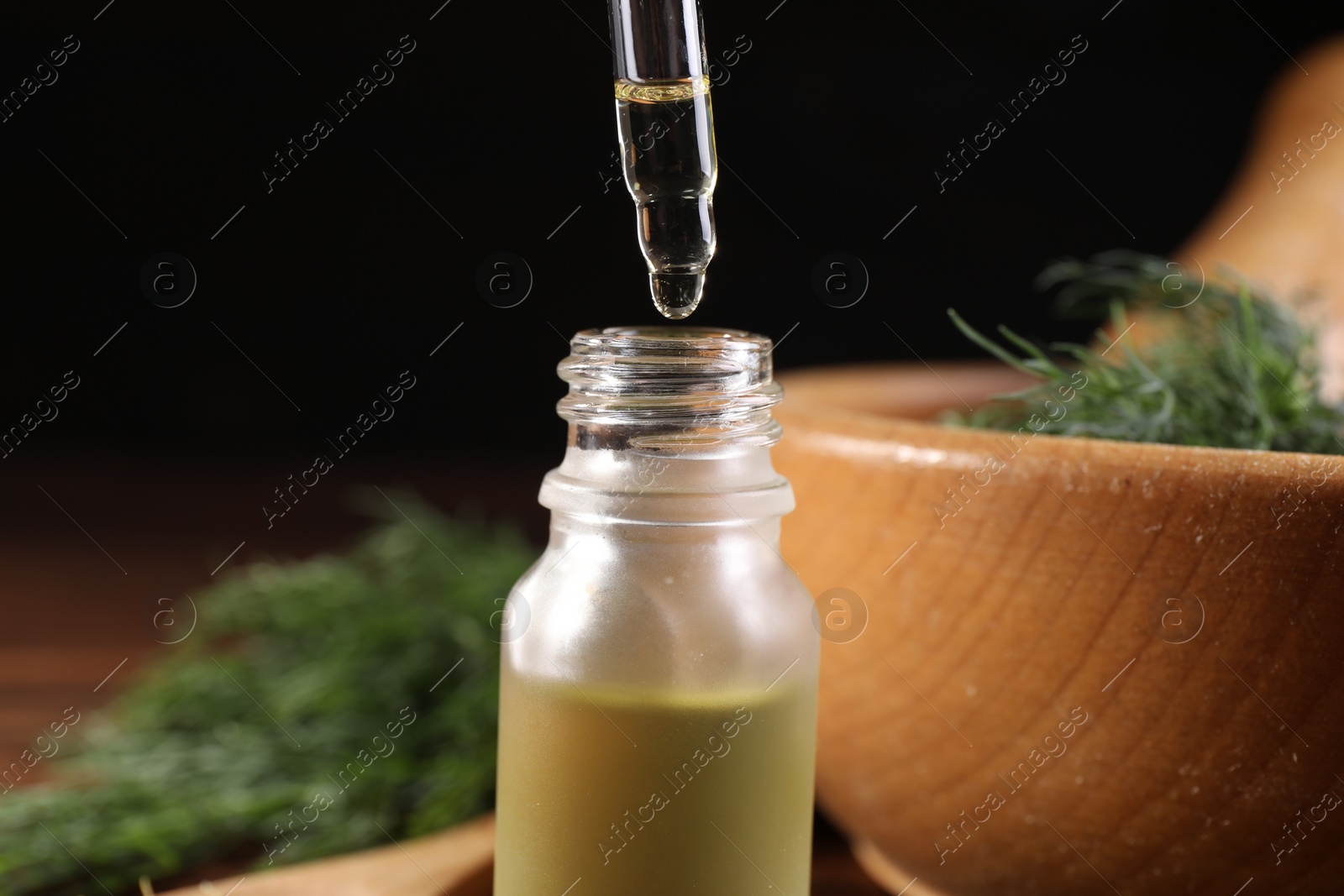 Photo of Dripping dill essential oil from pipette into bottle against black background, closeup