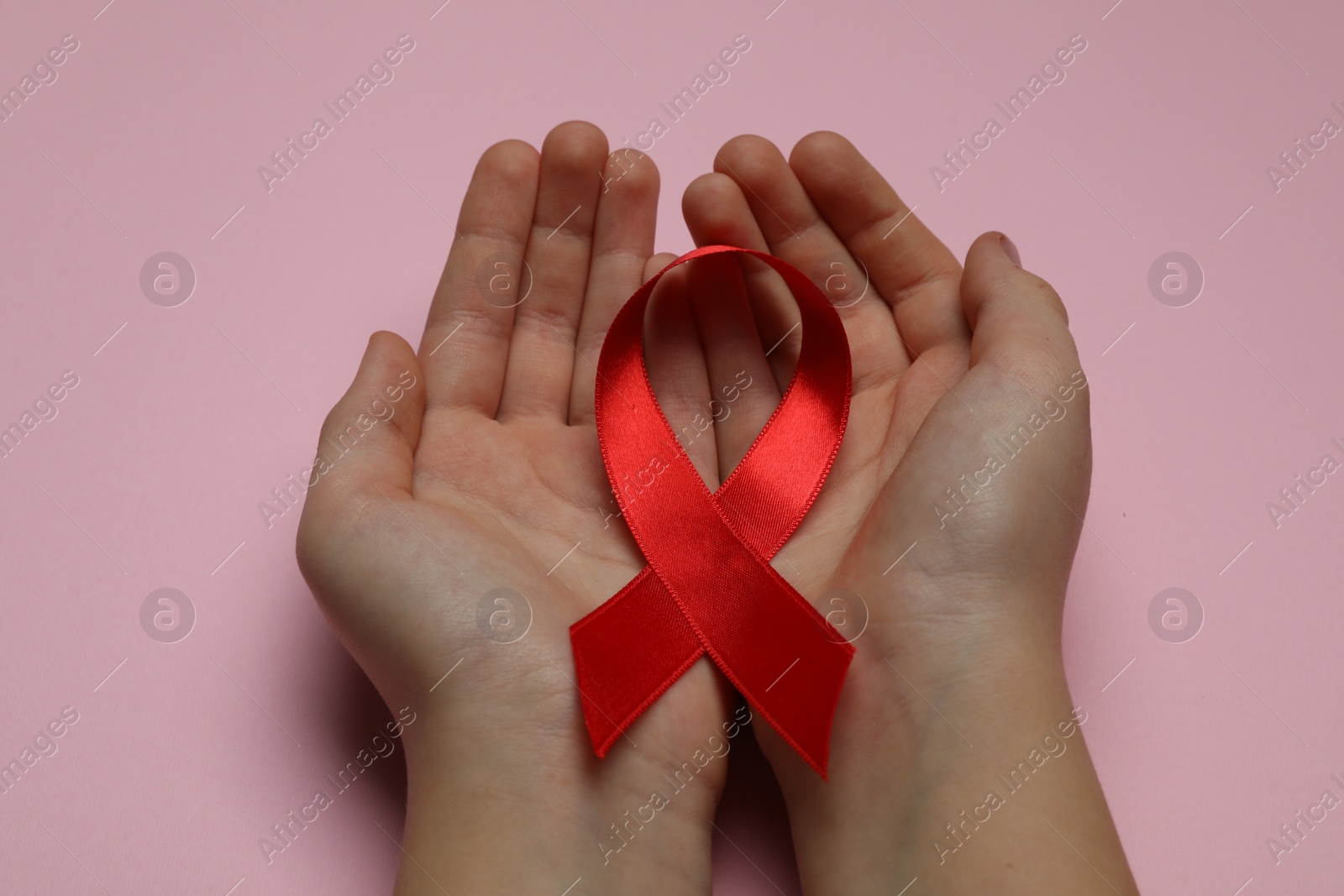 Photo of Little girl holding red ribbon on pink background, closeup. AIDS disease awareness