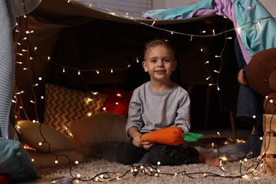 Photo of Happy boy with carrot toy in decorated play tent at home